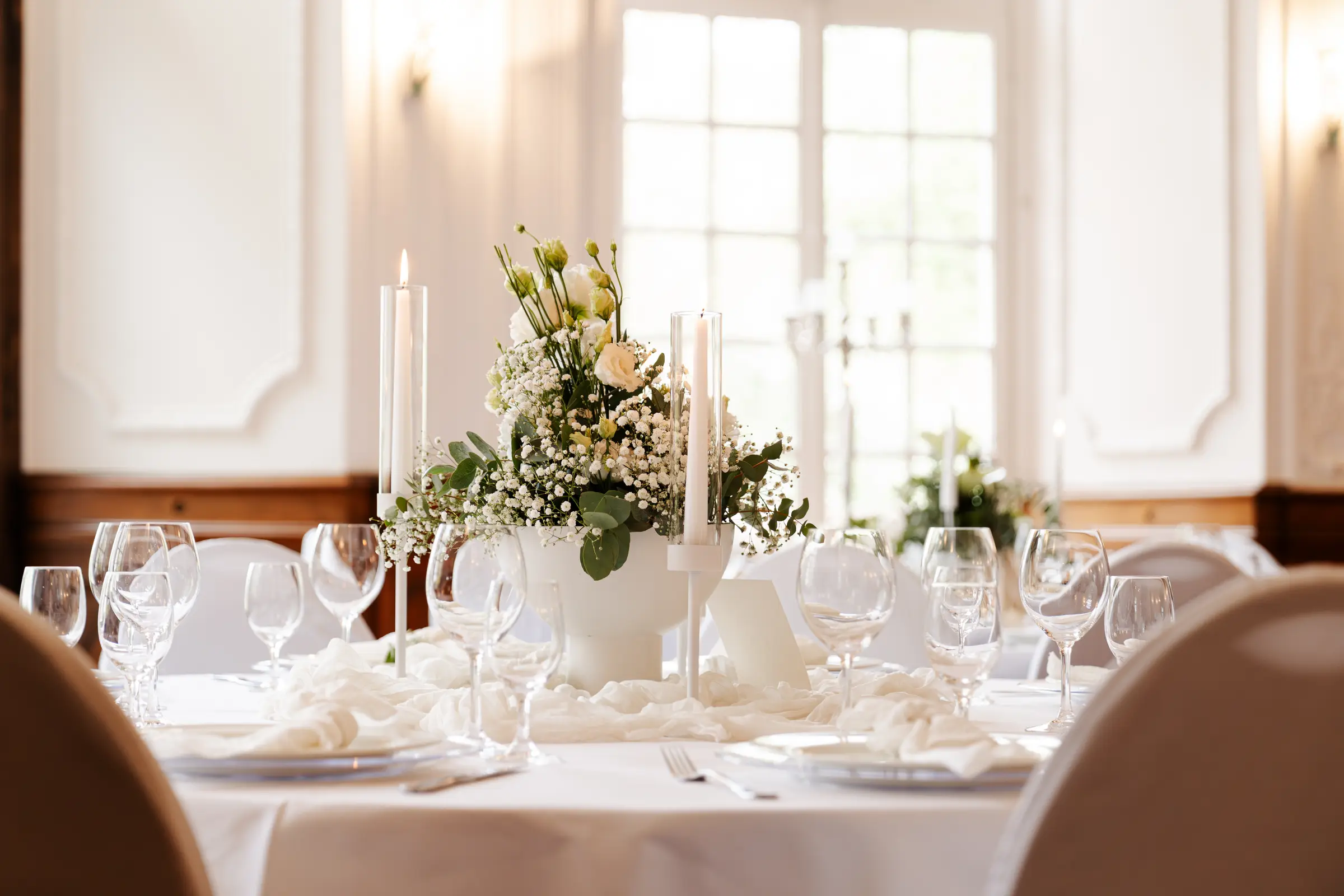 Festlich gedeckte Hochzeitstafel in einem hellen Saal: weißes Blumengesteck aus Rosen und Schleierkraut in einer Schale, weiße Stabkerzen, Weingläser und Chiffonläufer